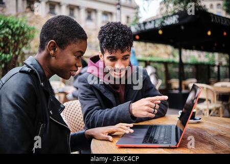 Man pointing toward laptop while sitting by friend at sidewalk cafe Stock Photo