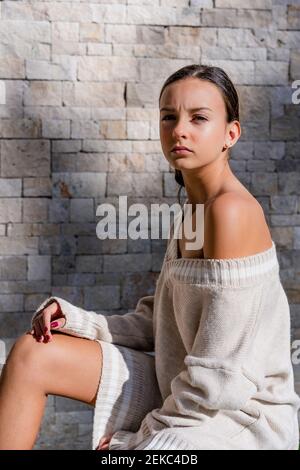 Stylish teenage girl wearing off shoulder dress posing by brick wall at home Stock Photo