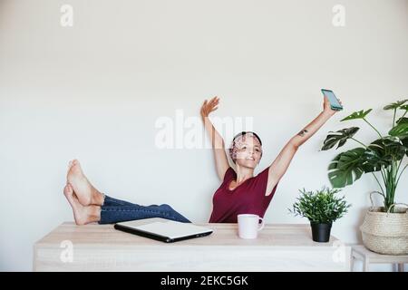 Relaxed businesswoman holding smart phone while looking away with feet up on table against wall Stock Photo