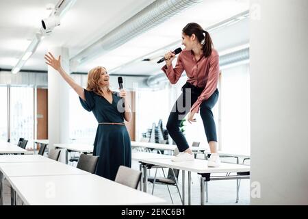Happy female colleagues singing on microphone at conference table while taking a break in office Stock Photo