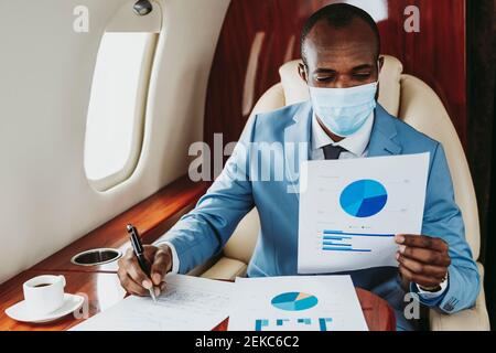 Young businessman analyzing graph while working in airplane during COVID-19 Stock Photo