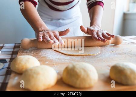 Woman flattening dough with rolling pin on cutting board to make croissants in kitchen Stock Photo