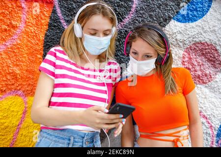 Female friends using mobile phone while listening music against colorful wall during COVID-19 Stock Photo