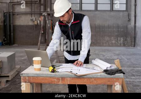 Male architect with blueprints using laptop on table while standing in building Stock Photo