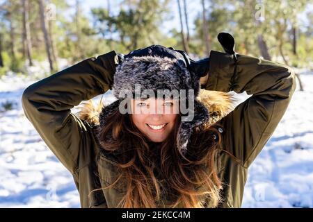 Smiling woman wearing fur cap standing with hands behind head at forest Stock Photo