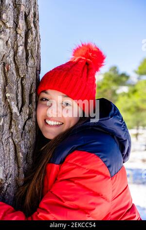 Young woman wearing red knit hat smiling while hugging tree standing in forest Stock Photo