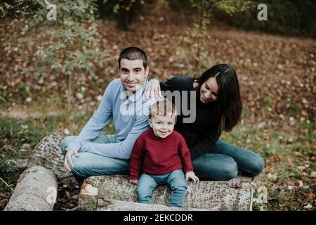 Smiling toddler boy staring while sitting on log in forest during autumn Stock Photo