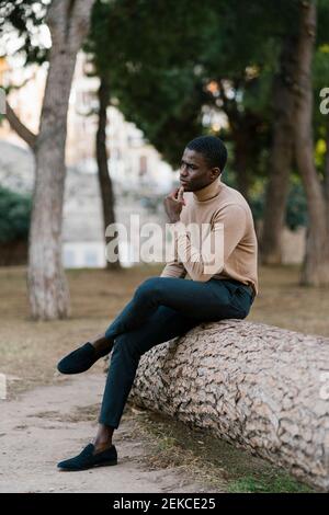 Thoughtful man looking away while sitting on fallen tree at park Stock Photo