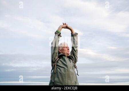 Senior male hiker looking away while stretching arms against cloudy sky Stock Photo