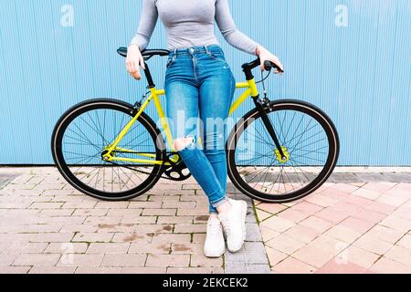Mid adult woman wearing torn jeans sitting on fixie bike against blue wall Stock Photo