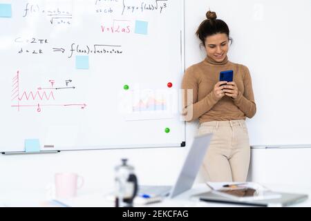 Smiling female student using smartphone while standing against wall Stock Photo