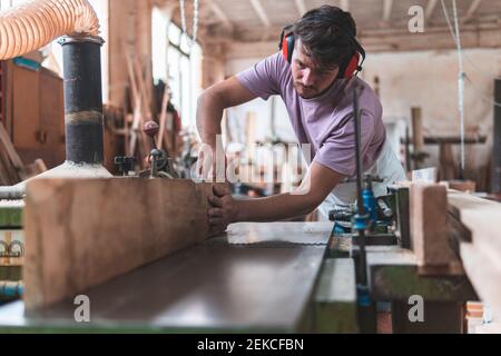 Young male craftsperson holding wooden plank on workbench while working in workshop Stock Photo