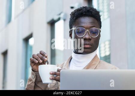 Young man stirring coffee while looking at laptop at sidewalk cafe Stock Photo