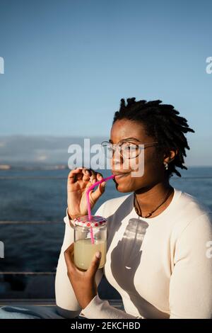 Young woman drinking juice while sitting against sky Stock Photo