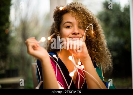 Smiling young woman with tangled Christmas lights on her sitting in living room Stock Photo