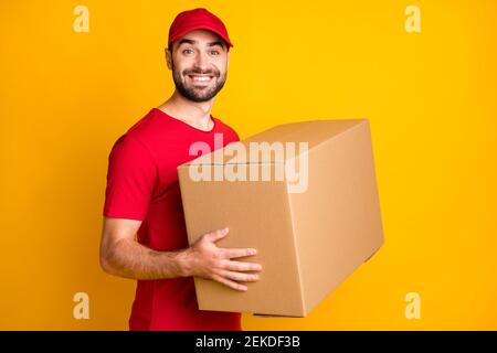Photo of cute delivery man wear red outfit smiling holding big brown box isolated yellow color background Stock Photo