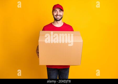 Photo of cheerful guy courier dressed red clothes delivering large brown box isolated yellow color background Stock Photo