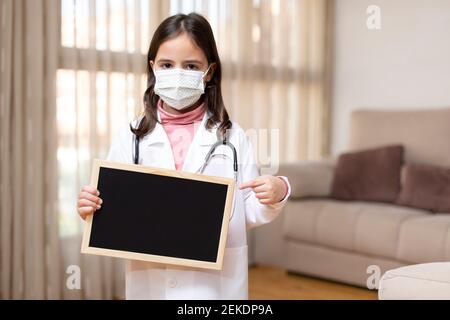 Little child dressed as a doctor and wearing a medical mask holding and pointing with her hand to a blackboard. She is at home. Space for text. Stock Photo