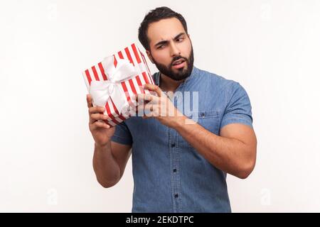 Curious bearded man shaking striped gift box trying to guess what is inside, wondered with present, surprises and bonuses. Indoor studio shot isolated Stock Photo