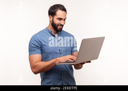 Smiling positive man holding laptop in hand and typing, blogger making posts in social networks, chatting with followers. Indoor studio shot isolated Stock Photo