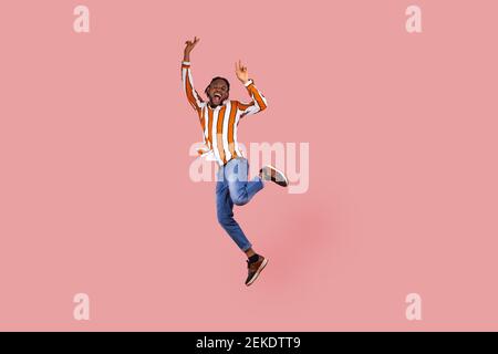 Full length excited ecstatic afro-american man with dreadlocks in striped shirt highly jumping posing, showing victory gesture, celebrating his triump Stock Photo
