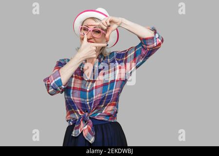 Portrait of attentive modern stylish mature woman in casual style with hat and eyeglasses standing with crop composition gesture and looking at target Stock Photo