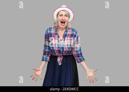 Portrait of nervous aggressive stylish mature woman in casual style with hat standing, raised arms with angry face and looking at camera screaming. in Stock Photo