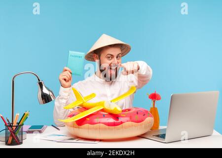 Happy relaxed man with beard in funny hat pointing finger at you holding passport recommending hot tour abroad, lifebuoy and toy plane lying on workpl Stock Photo