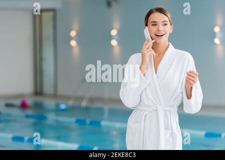 excited young woman in white bathrobe talking on smartphone in spa center Stock Photo