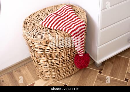 One hat with red and white stripes with a big red pompom is on the  wicker basket next to a white wall and white furniture. Stock Photo