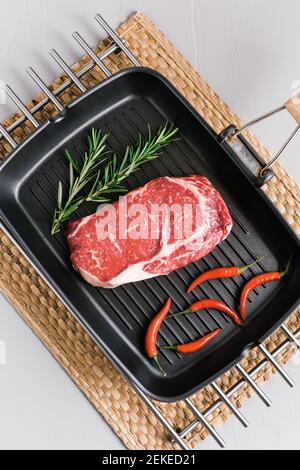 Raw rib eye beef steak on black grilling pan with chili pepper and rosemary, top view. Stock Photo