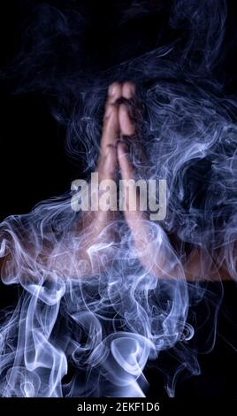 a out of focused human folded hands surrounded by blue smoke in black background Stock Photo