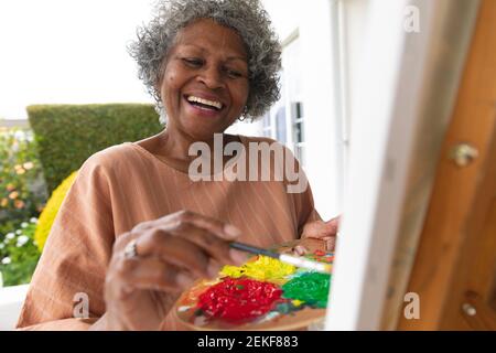 African american senior woman smiling while painting on canvas standing on porch of the house Stock Photo