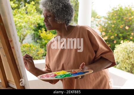 Thoughtful african american senior woman painting on canvas while standing on porch of the house Stock Photo