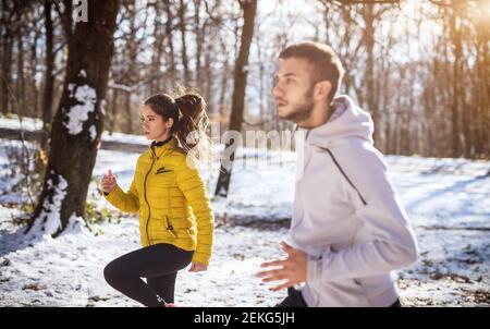 Close up side view of young focused attractive beautiful fitness sporty couple in winter sportswear warming in the snowy forest. Stock Photo