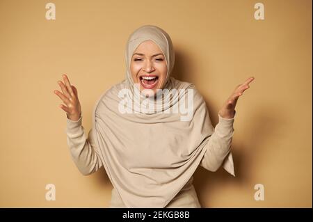 Arab Muslim woman in hijab poses in front of the camera against a beige background, expressing anger and irritation by shouting. Copy space. Strict fo Stock Photo