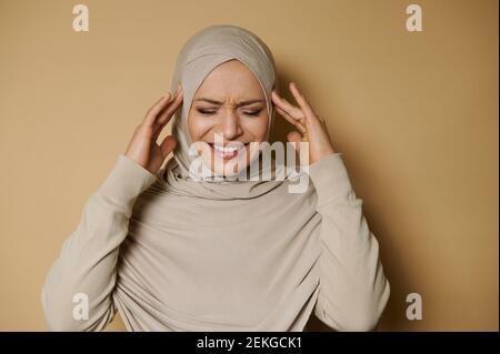 Beautiful arab woman in hijab posing to the camera expressing pain and headache on a beige background with copy space. Strict formal outfit Stock Photo