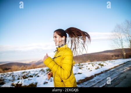 Young active athletic girl jogging in winter sportswear on snowy winter road with earphones in the sunny morning. Stock Photo