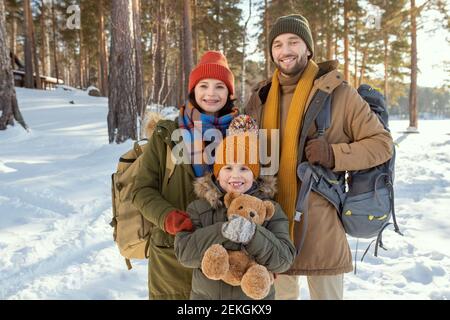 Cheerful young family of three in warm winterwear standing in front of camera against natural environment and looking at you outside Stock Photo