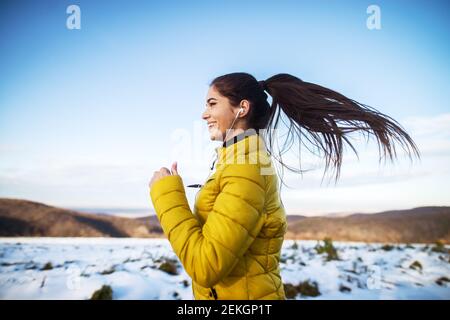 Young active athletic girl jogging in winter sportswear on snowy winter road with earphones in the sunny morning. Stock Photo