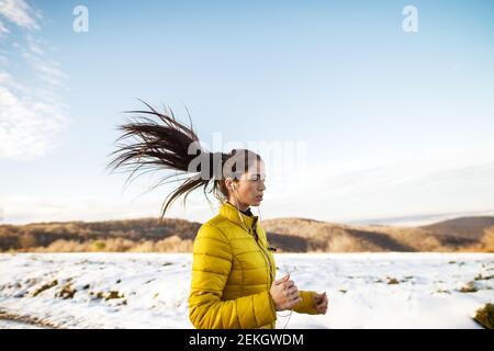 Young active athletic girl jogging in winter sportswear on snowy winter road with earphones in the sunny morning. Stock Photo