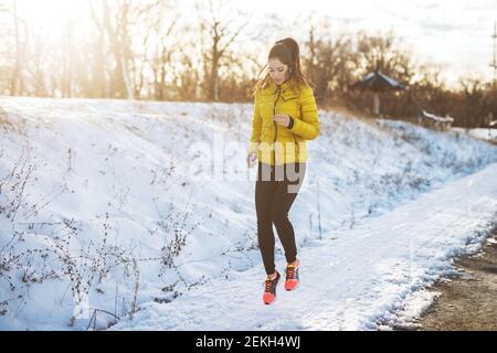 Young active athletic girl jogging in winter sportswear on snowy winter road with earphones in the sunny morning. Stock Photo