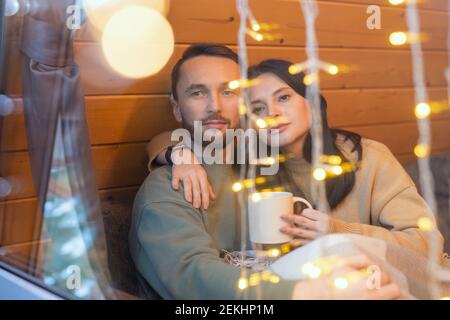 Young serene affectionate couple in sweaters sitting on couch by large window of country house on winter weekend and enjoying rest Stock Photo