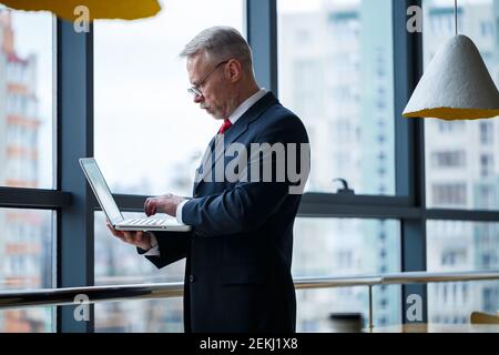 Smiling happy managing director thinks about his successful career development while standing with a laptop in his office near the background of a win Stock Photo