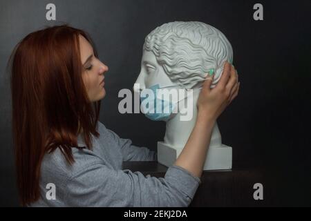 ginger young woman girl on a black background a gray t-shirt and plaster bust in protective medical mask Stock Photo