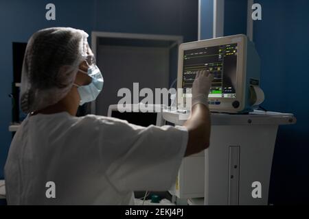 Young female assistant in gloves, mask and uniform pointing at display of medical equipment while taking control of patient body condition Stock Photo