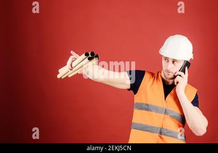 Supply of building materials concept. Engineer, architect on busy face speaks on phone, holds blueprints, pointing with finger, copy space. Man supervises construction on phone, red background. Stock Photo