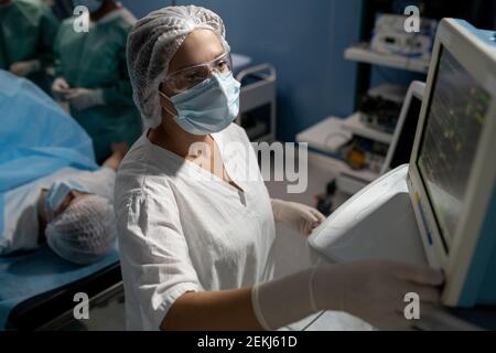 Serious female assistant of surgeon in gloves, mask and uniform looking at display of medical equipment against patient before operation Stock Photo