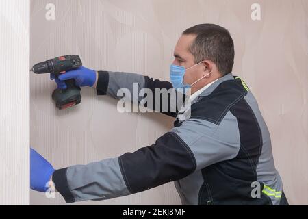 Repairman in overalls works as screwdriver. Master wearing  medical mask and gloves complies with safety rules during pandemic. Stock Photo