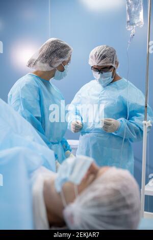 Young female assistant and male surgeon in protective workwear preparing medical instruments before operation with patient in front Stock Photo